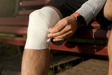 Photo of Man applying bandage onto his knee on wooden bench outdoors, closeup