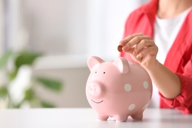 Woman putting coin into piggy bank at table indoors, closeup. Space for text