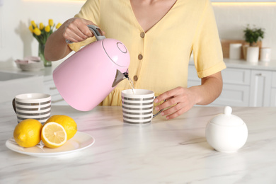 Photo of Woman pouring hot water from modern electric kettle in cup indoors, closeup