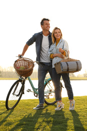 Young couple with bicycle and picnic basket near lake on sunny day