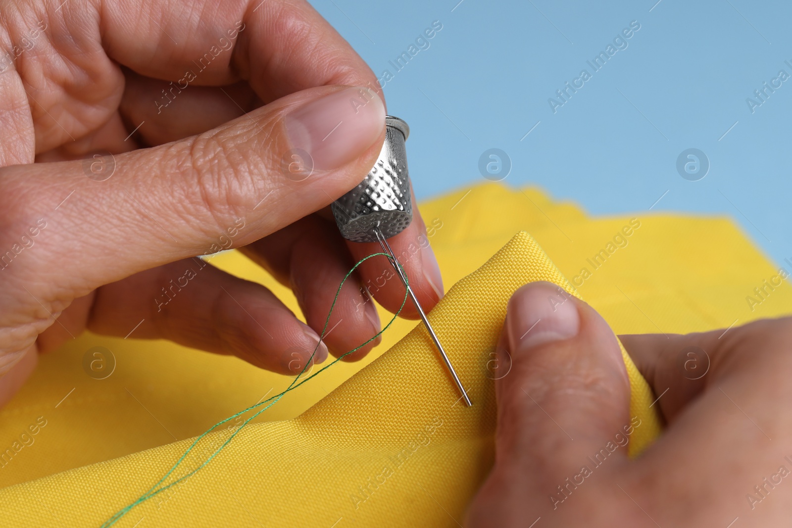 Photo of Woman sewing on yellow cloth with thimble and needle against light blue background, closeup