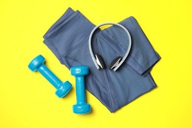 Photo of Stylish sports leggings, headphones and dumbbells on yellow background, flat lay