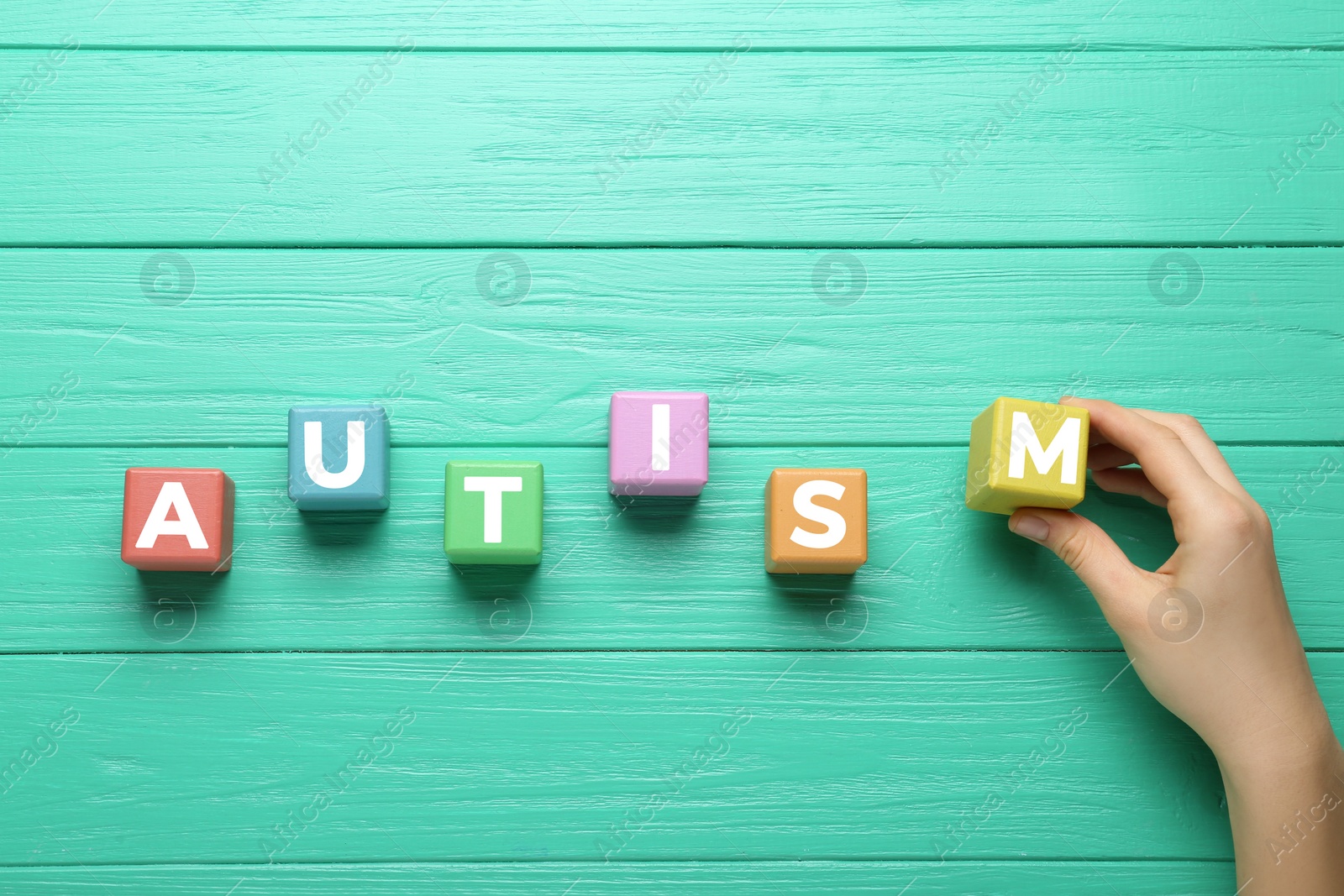 Photo of Woman making word Autism with colorful cubes on turquoise wooden table, top view