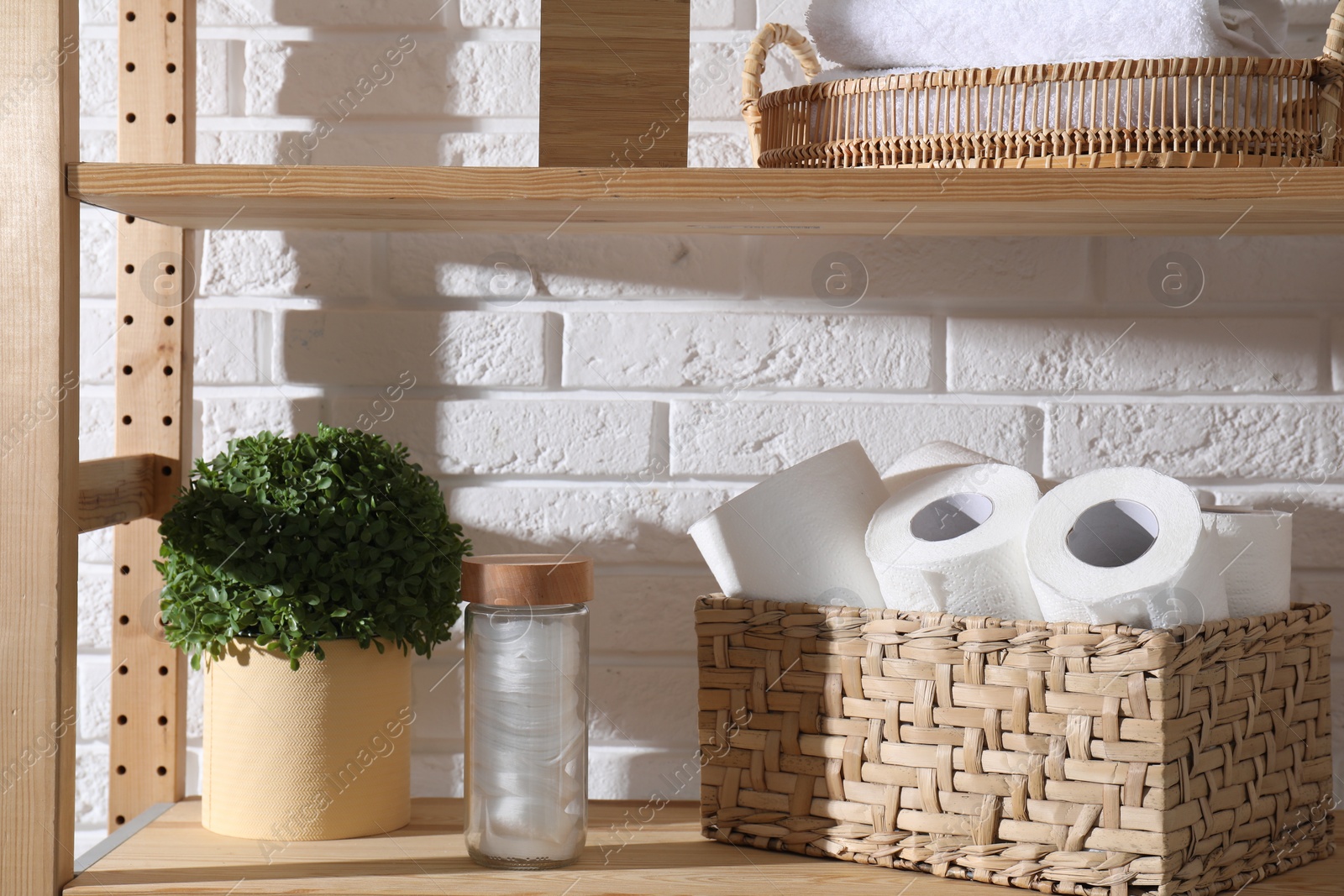 Photo of Toilet paper rolls in wicker basket, floral decor and cotton pads on wooden shelf against white brick wall