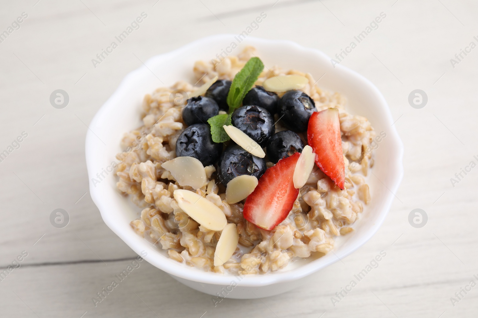 Photo of Tasty oatmeal with strawberries, blueberries and almond petals in bowl on white wooden table, closeup