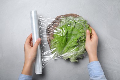 Photo of Woman putting plastic food wrap over fresh lettuce at light grey table, top view