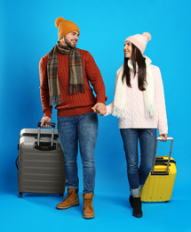 Happy young couple in warm clothes with suitcases on blue background. Winter vacation