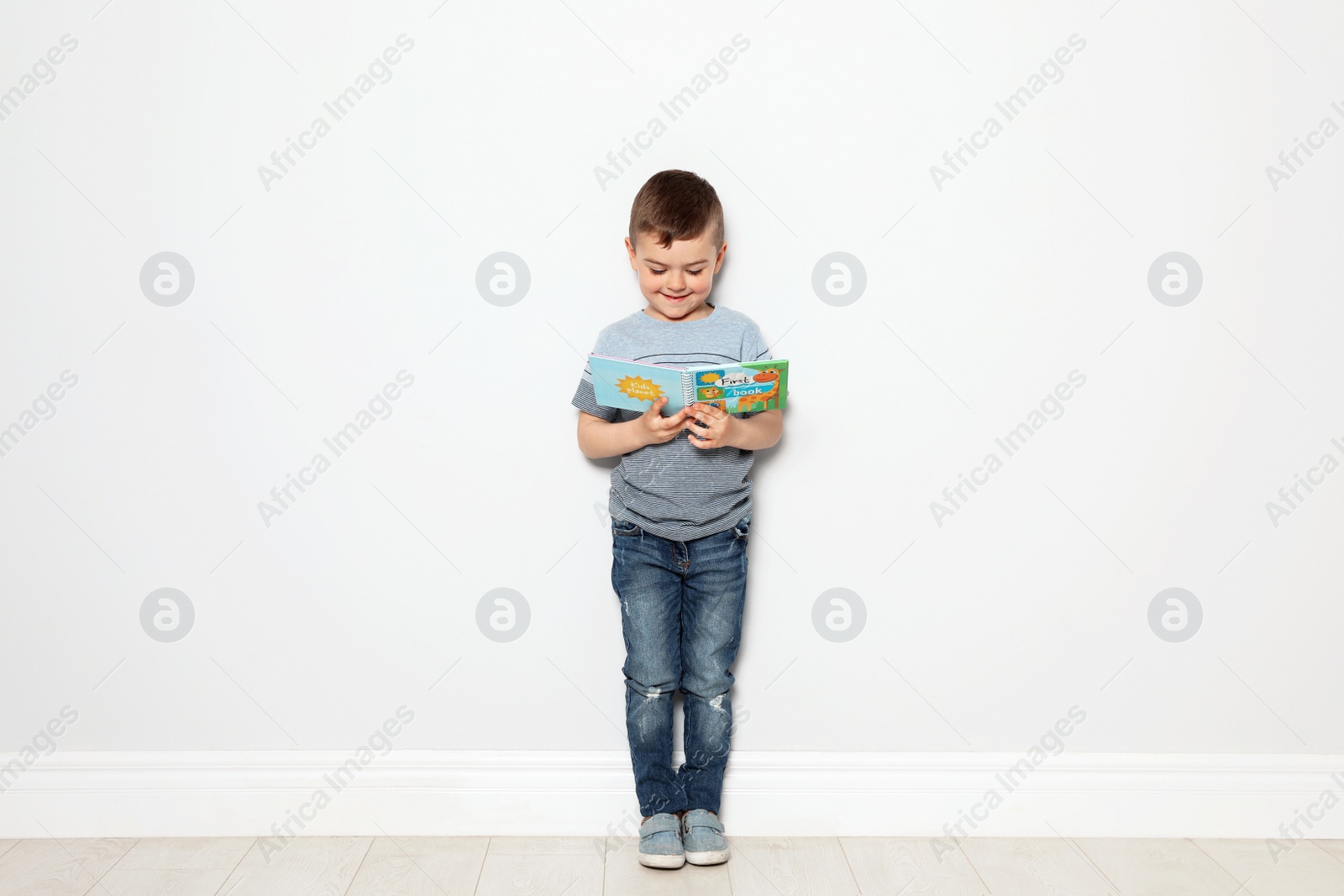 Photo of Cute little boy reading book near white wall, space for text