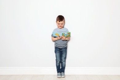 Photo of Cute little boy reading book near white wall, space for text