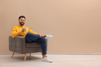 Handsome man with cup of drink sitting in armchair near beige wall indoors, space for text