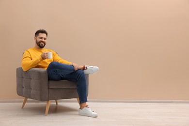 Photo of Handsome man with cup of drink sitting in armchair near beige wall indoors, space for text