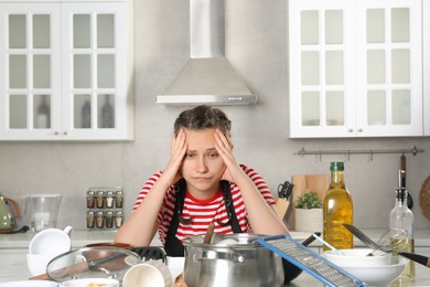 Upset woman in messy kitchen. Many dirty dishware and utensils on table