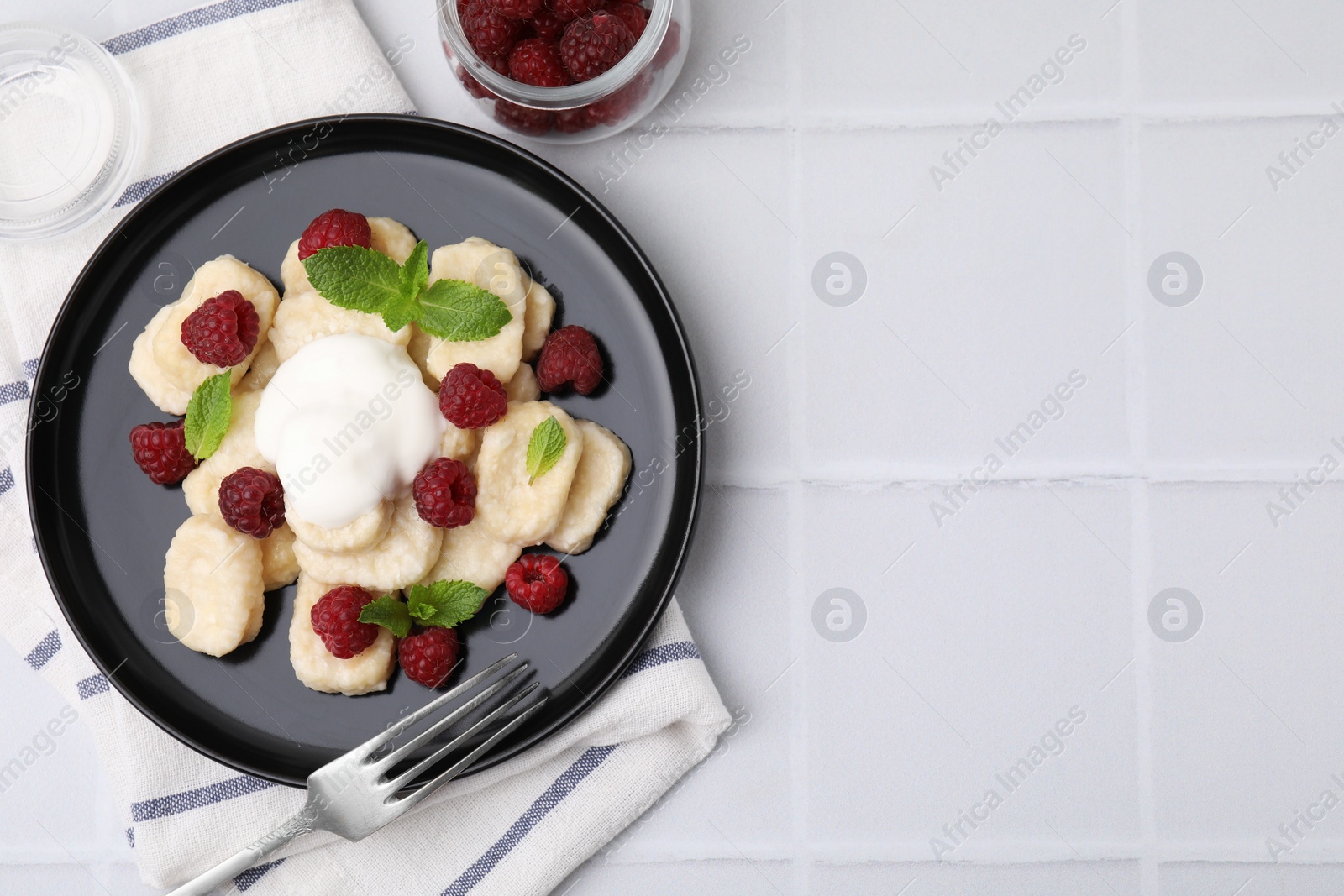 Photo of Plate of tasty lazy dumplings with raspberries, sour cream and mint leaves on white tiled table, flat lay. Space for text