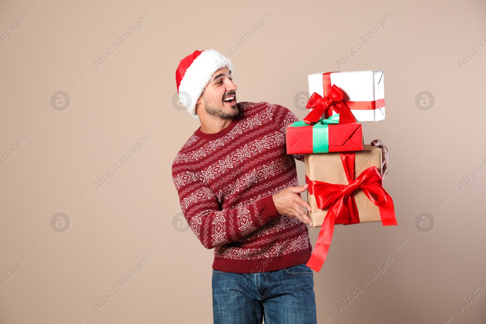 Photo of Happy man in Christmas sweater and Santa hat holding gift boxes on beige background