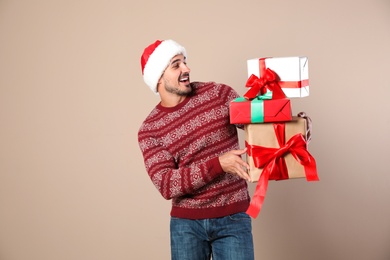 Happy man in Christmas sweater and Santa hat holding gift boxes on beige background