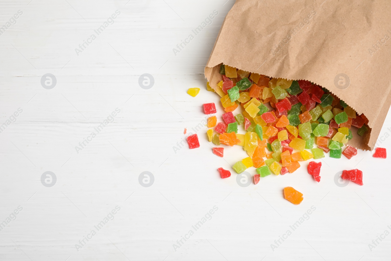 Photo of Overturned paper bag with mix of delicious candied fruits on white wooden table, flat lay. Space for text