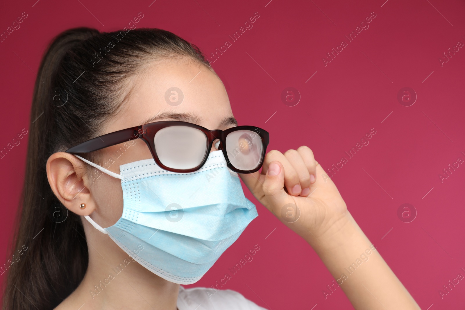 Photo of Little girl wiping foggy glasses caused by wearing medical face mask on pink background. Protective measure during coronavirus pandemic