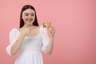 Happy woman pointing at credit card on pink background, space for text