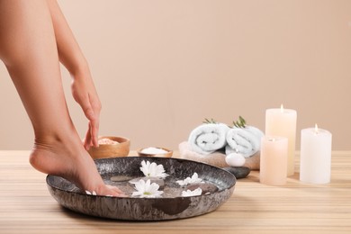 Photo of Woman soaking her foot in bowl with water and chrysanthemum flowers on wooden surface, closeup. Pedicure procedure
