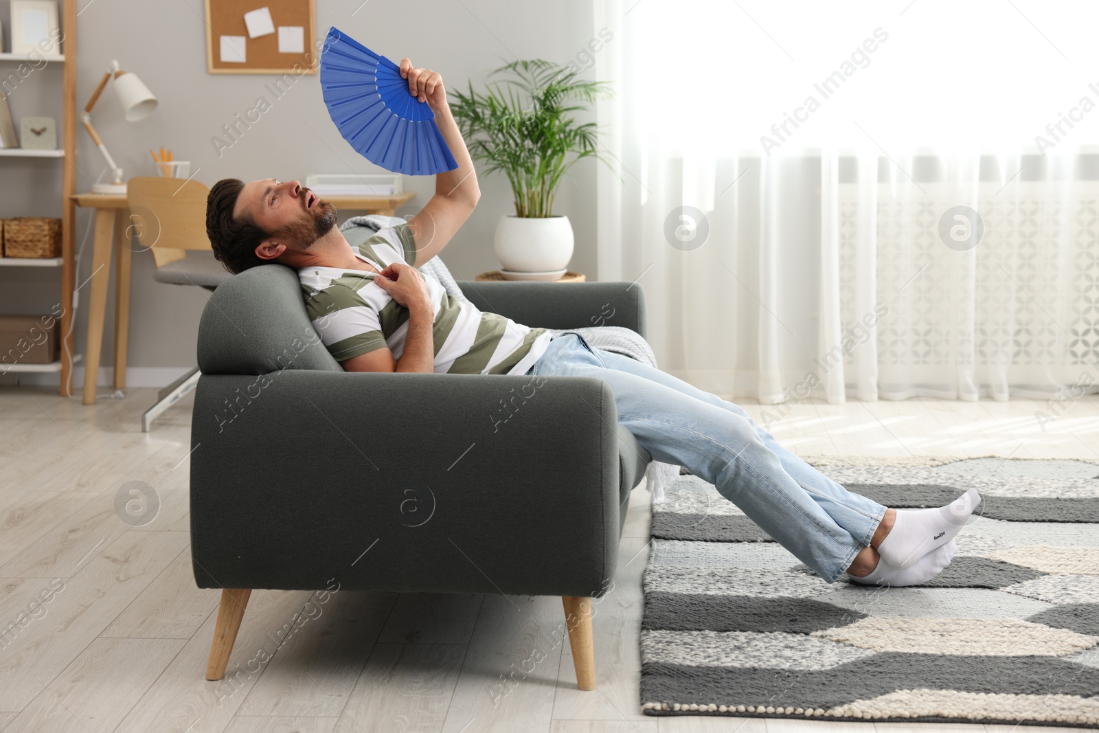 Photo of Bearded man waving blue hand fan to cool himself on sofa at home