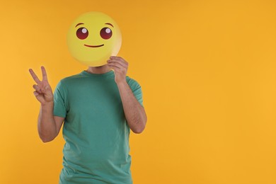 Photo of Man covering face with smiling emoticon and showing peace sign on yellow background. Space for text