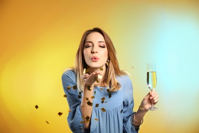 Portrait of happy woman with champagne in glass blowing off confetti on color background