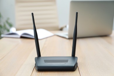 Photo of Modern Wi-Fi router on wooden table with laptop indoors