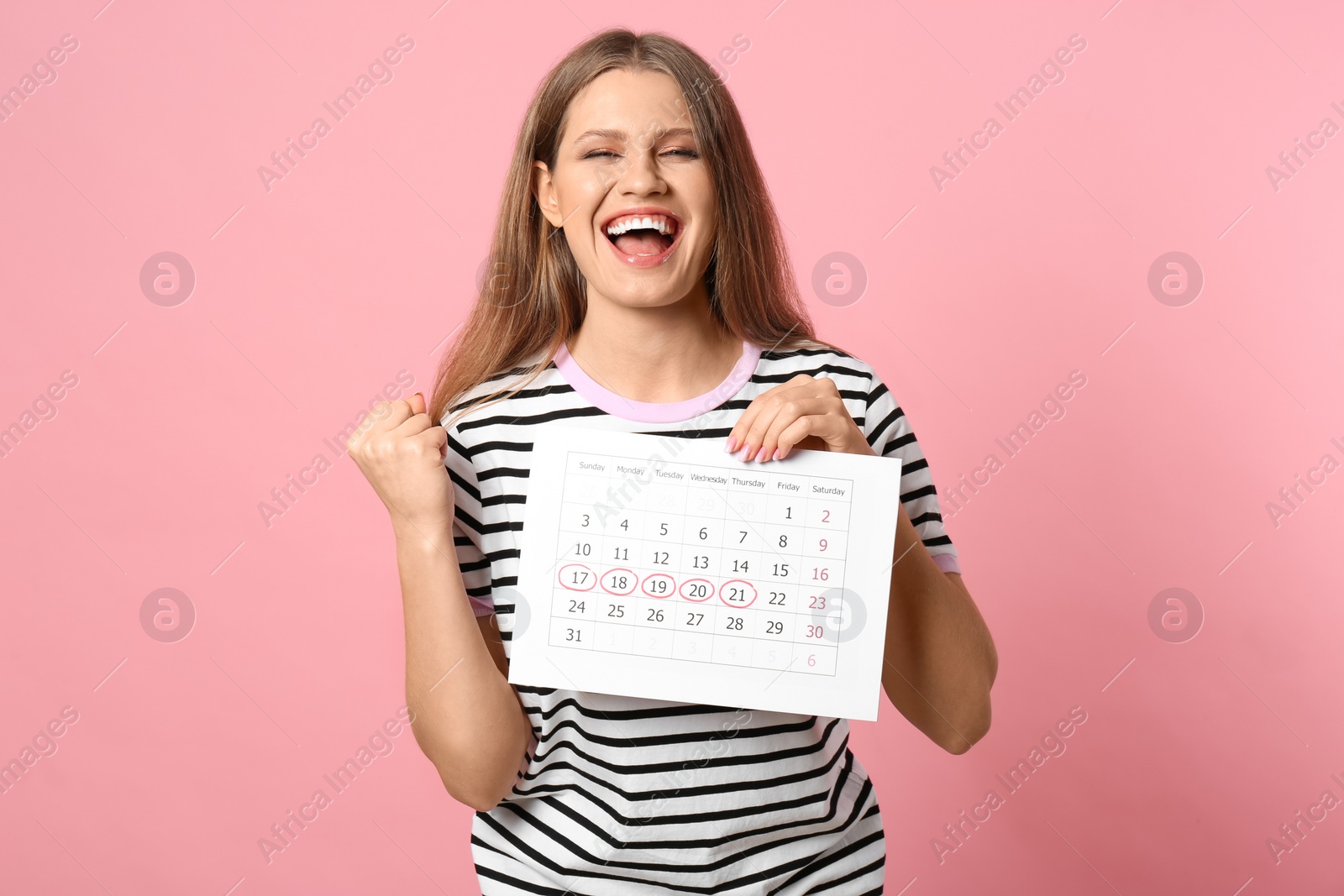 Photo of Excited young woman holding calendar with marked menstrual cycle days on pink background