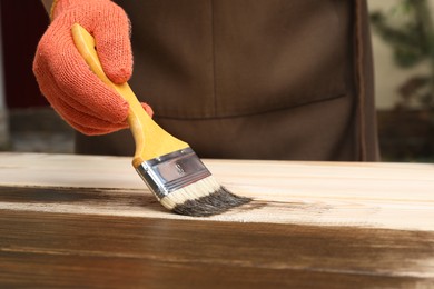 Photo of Man applying wood stain onto wooden surface against blurred background, closeup