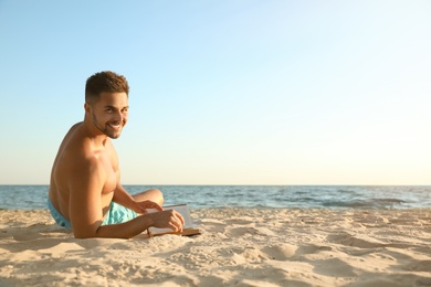 Young man reading book on sandy beach near sea