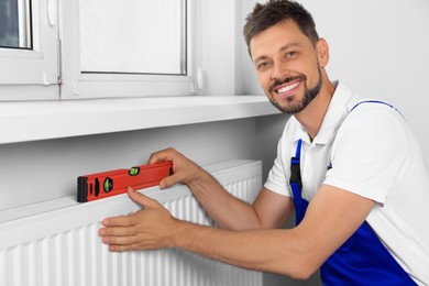 Professional plumber using bubble level for installing new heating radiator in room