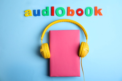 Tome with headphones and word AUDIOBOOK on light blue background, flat lay