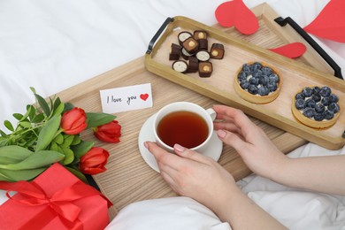 Photo of Tasty breakfast served in bed. Woman with tea, desserts, gift box, flowers and I Love You card at home, closeup