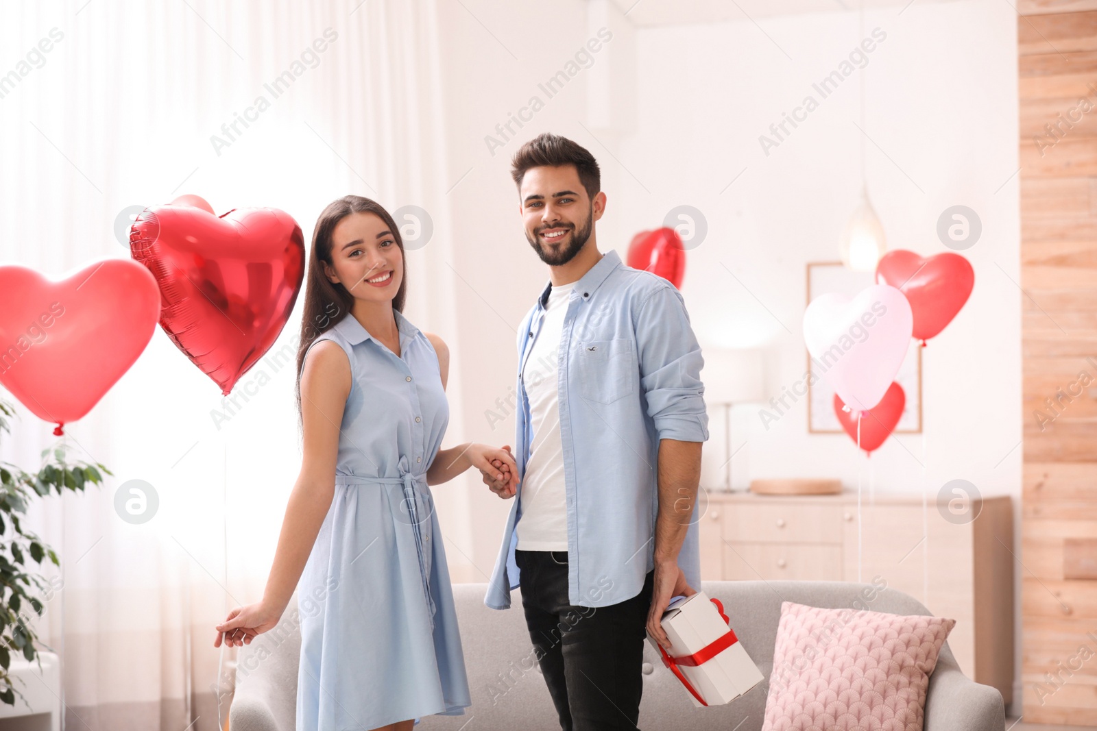 Photo of Young man presenting gift to his girlfriend in living room decorated with heart shaped balloons. Valentine's day celebration