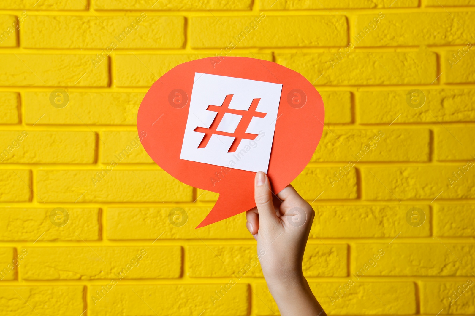 Photo of Woman holding red paper speech bubble with hashtag symbol against yellow brick wall, closeup