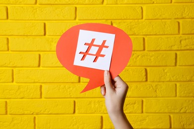 Photo of Woman holding red paper speech bubble with hashtag symbol against yellow brick wall, closeup