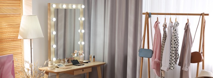 Image of Makeup room. Stylish dressing table with mirror, chair and clothing rack indoors, banner design