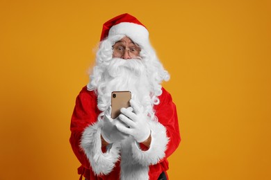 Photo of Merry Christmas. Santa Claus using smartphone on orange background