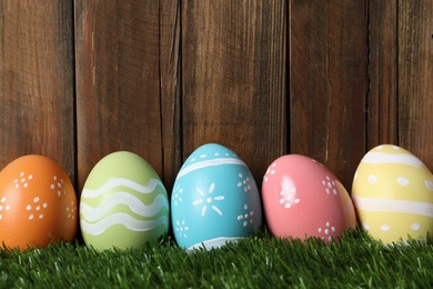 Colorful painted Easter eggs on green grass against wooden background, space for text