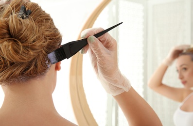 Photo of Young woman applying hair dye on roots in front of mirror at home, closeup