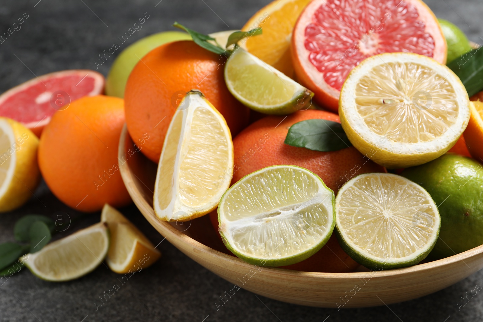 Photo of Different fresh citrus fruits and leaves in bowl on grey textured table, closeup