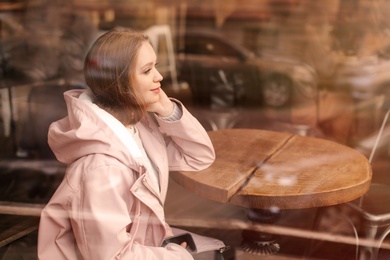 Photo of Beautiful young woman sitting at table in cafe, view from outdoors through window