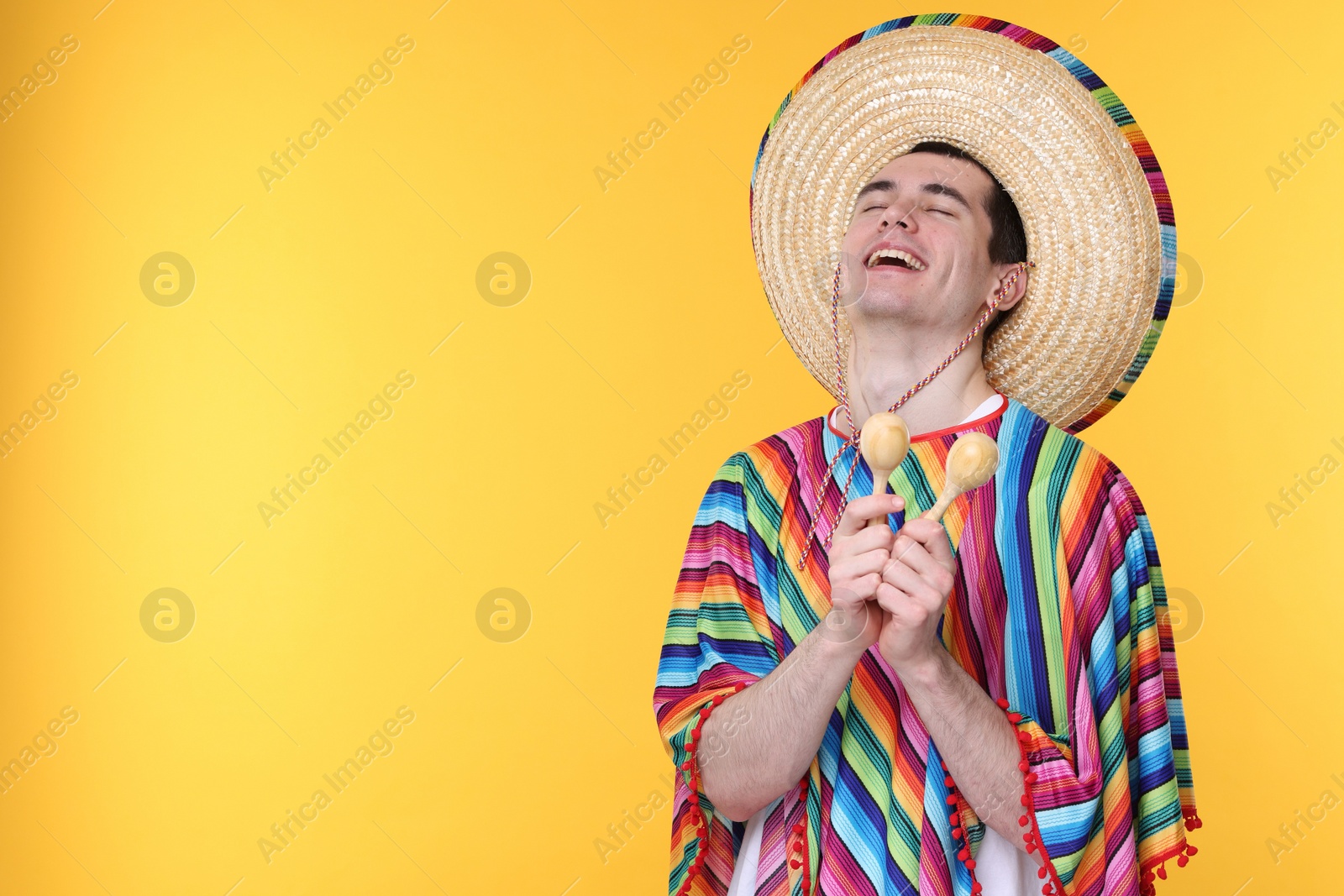 Photo of Young man in Mexican sombrero hat and poncho with maracas on yellow background. Space for text