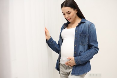 Photo of Pregnant young woman touching her belly near window indoors