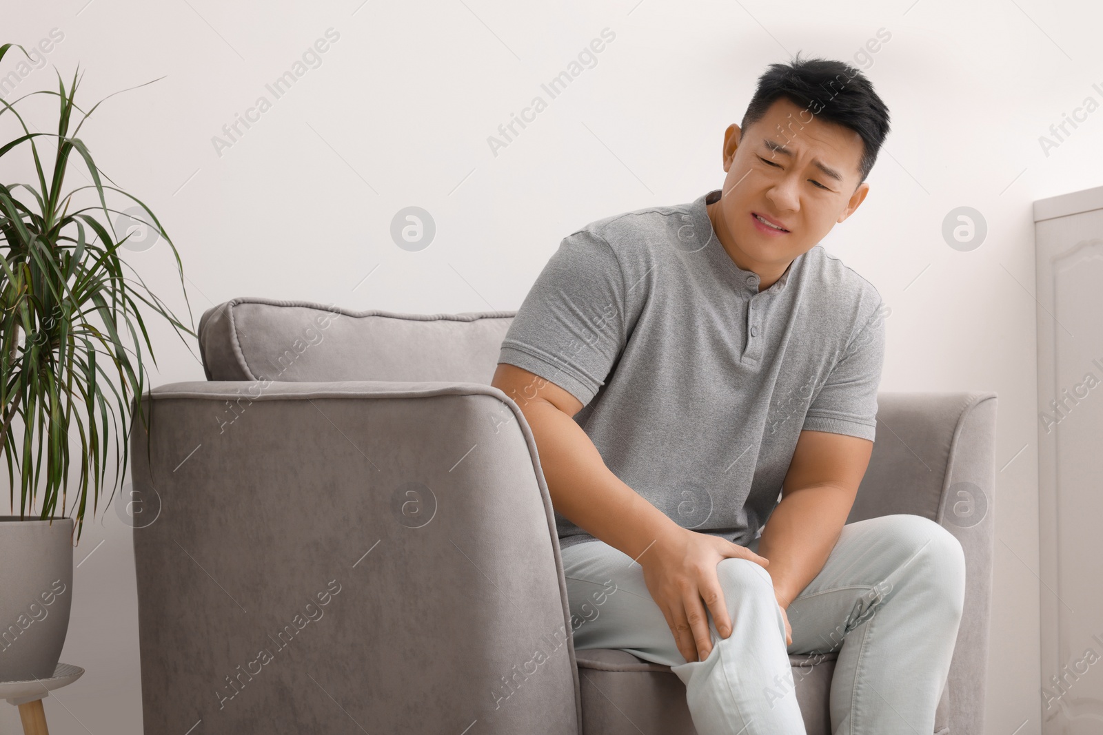 Photo of Asian man suffering from knee pain on armchair indoors