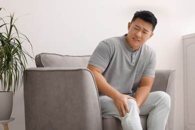 Photo of Asian man suffering from knee pain on armchair indoors