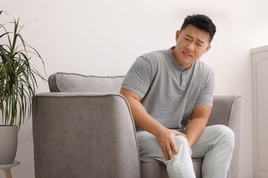 Asian man suffering from knee pain on armchair indoors