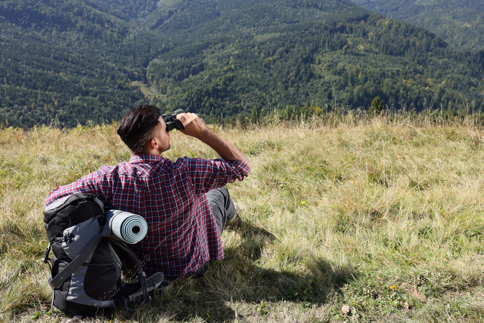 Photo of Tourist with hiking equipment looking through binoculars in mountains, back view