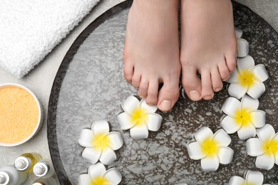 Photo of Woman soaking her feet in bowl with water and flowers on light grey floor, top view. Spa treatment