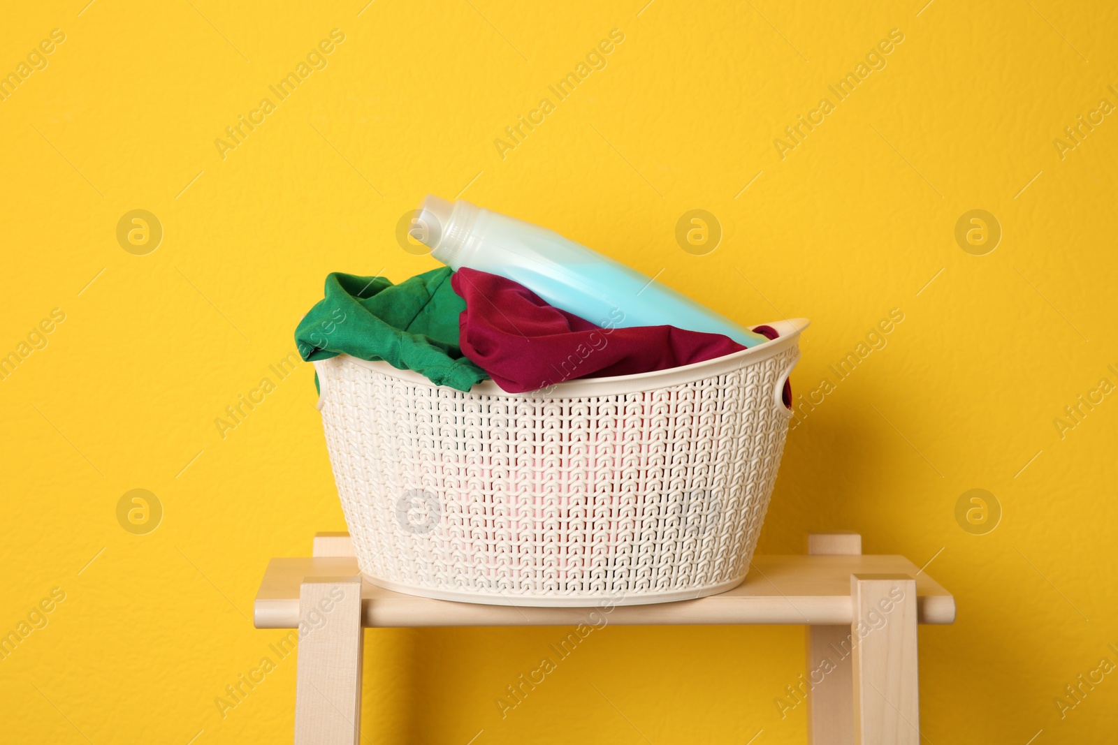 Photo of Laundry basket with dirty clothes and detergent on wooden stool against yellow background